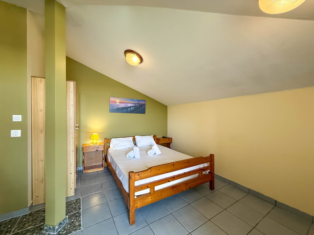 A budget double room features a spacious bed with neatly arranged linens and two towels placed on top. Warm yellow and green walls introduce a calming atmosphere. A small wooden bedside table and lamp accompany the bed, while a door leads to additional space.