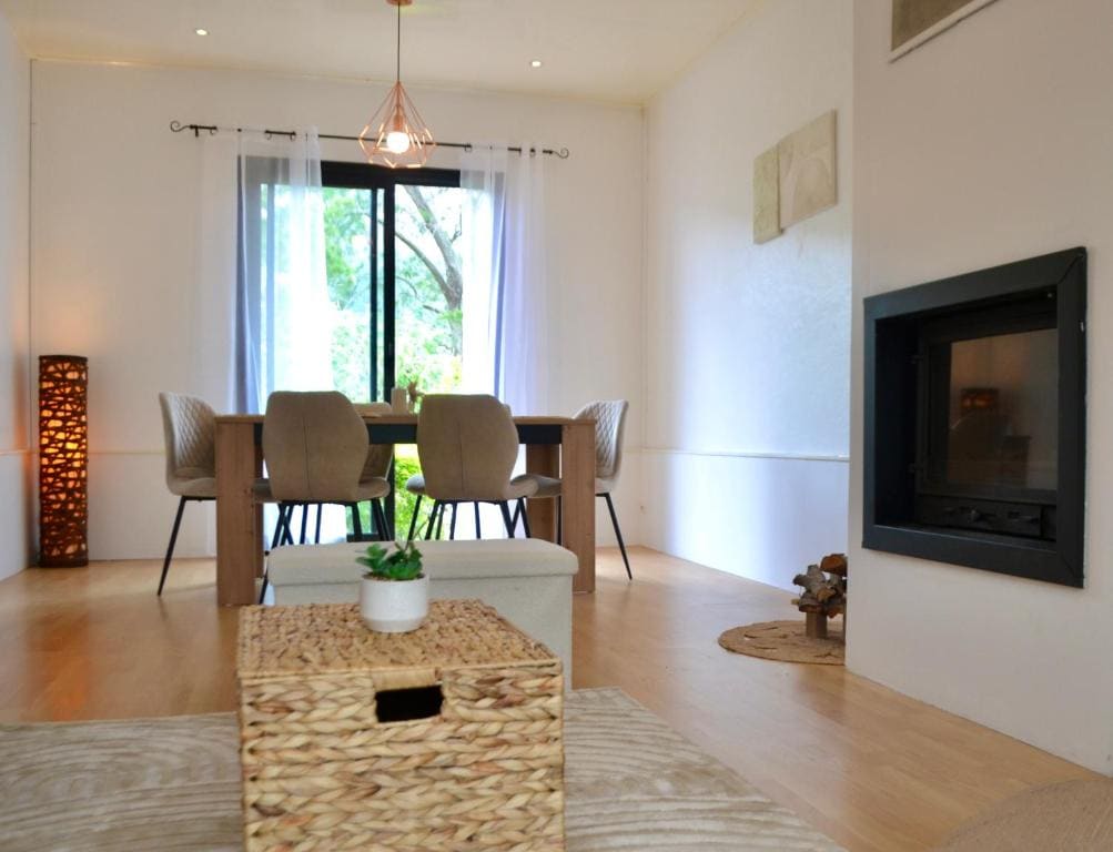 An inviting living area showcases a modern dining table surrounded by four upholstered chairs. Natural light filters through a large window, enhancing the neutral color palette. A cozy fireplace is positioned against one wall, while a woven storage basket and decorative elements add texture to the space.