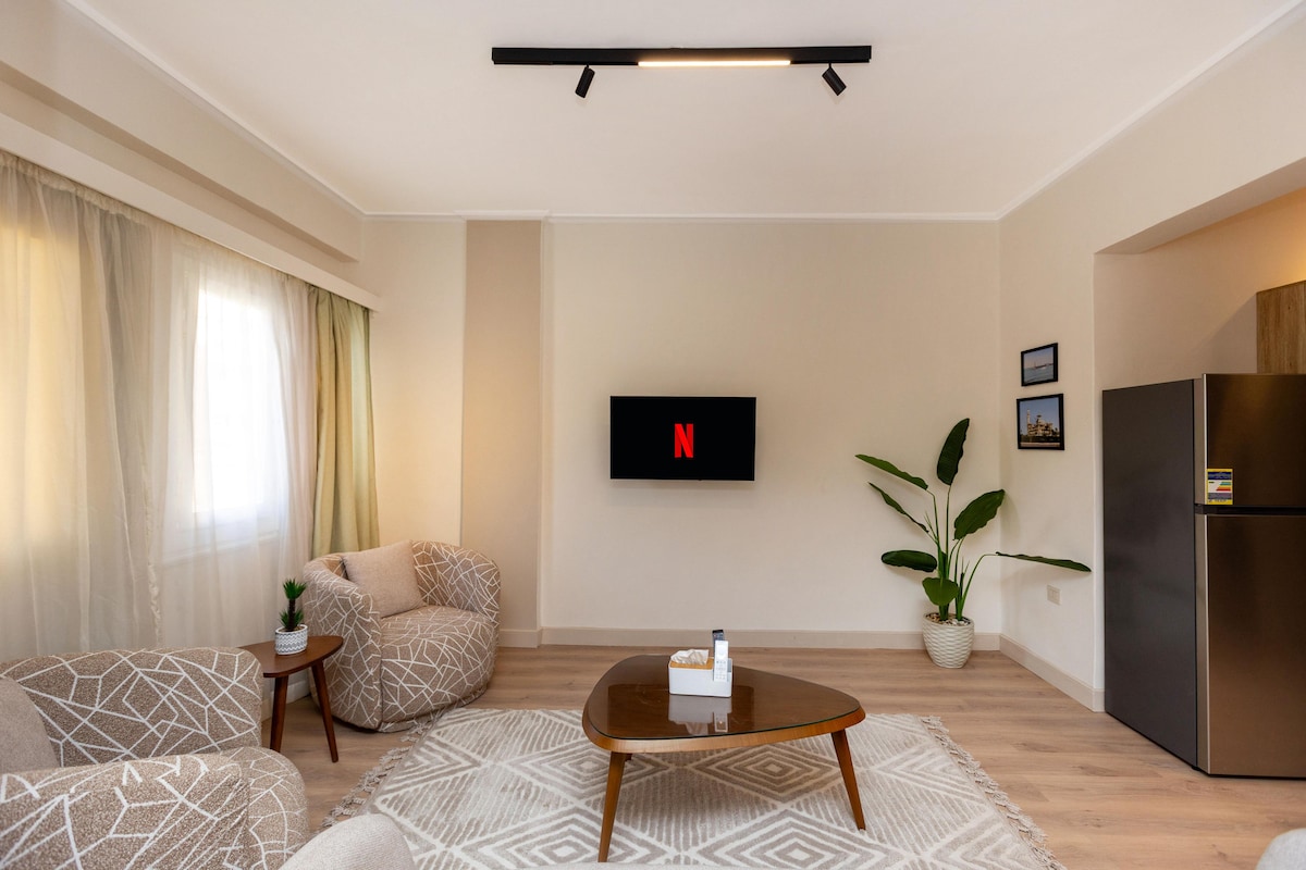 A bright living area features a modern design with a soft, neutral color palette. A cozy seating arrangement includes patterned chairs and a low coffee table, all situated under a beam of light. A television is mounted on the wall, with a small plant adding a touch of greenery.
