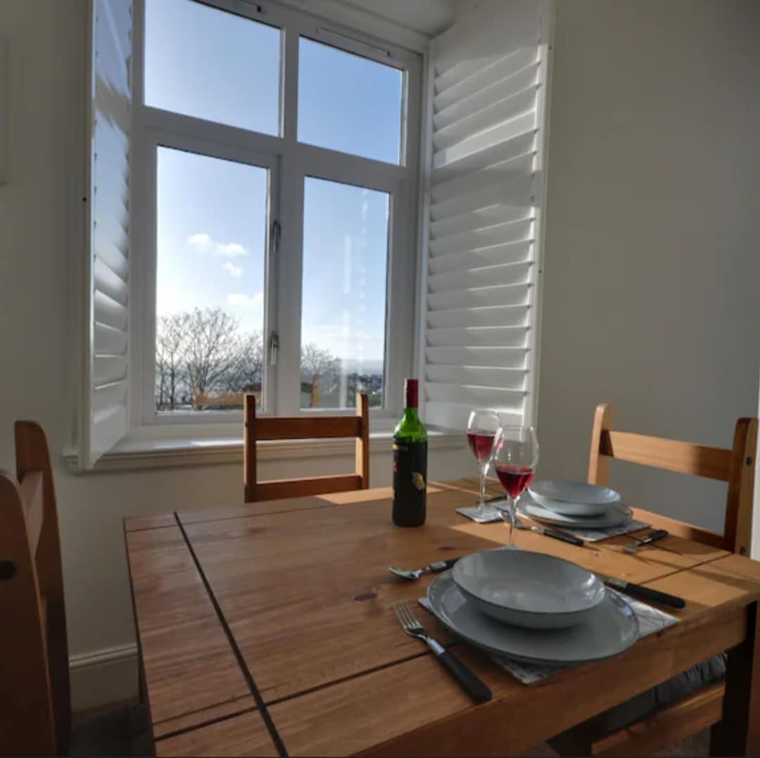 A dining table set for two is visible, featuring plates, wine glasses, and cutlery. The light wood table is positioned near a large window with shutters, providing a view of the outdoors. Bright natural light fills the space, creating a welcoming ambiance.
