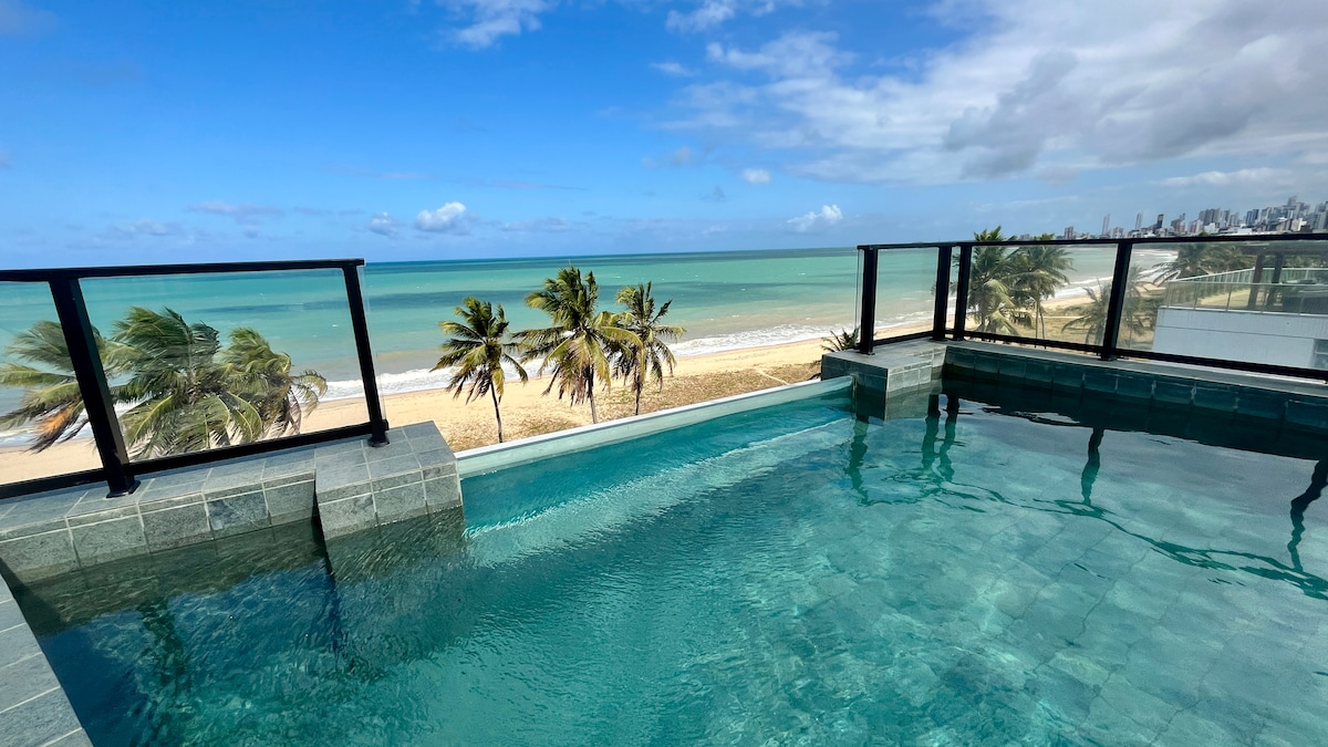 A rooftop pool reflects the clear blue sky and ocean, featuring a modern design with stone accents. The surrounding area is bordered by black railings, offering unobstructed views of the sandy beach and palm trees swaying with the breeze.