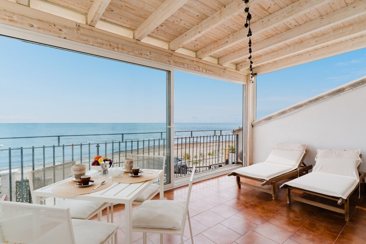 A bright seating area features a table set for two, with a view of the sea visible through expansive glass doors. Two loungers are positioned nearby, showcasing a spacious and inviting design. Natural light fills the room, enhancing the coastal ambiance.