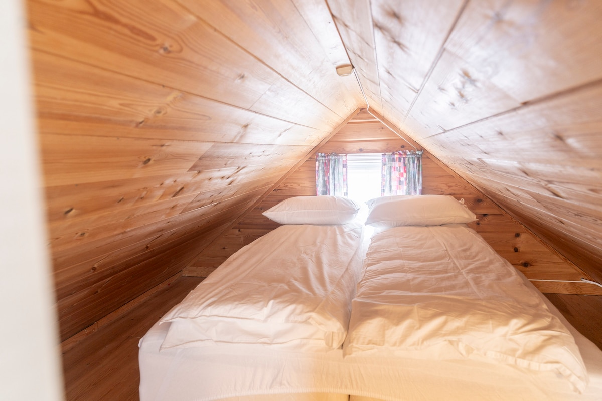 A lofted sleeping area features a double bed with white bedding against warm wooden walls. Large windows adorned with colorful curtains allow natural light to brighten the space, creating a cozy and inviting environment.