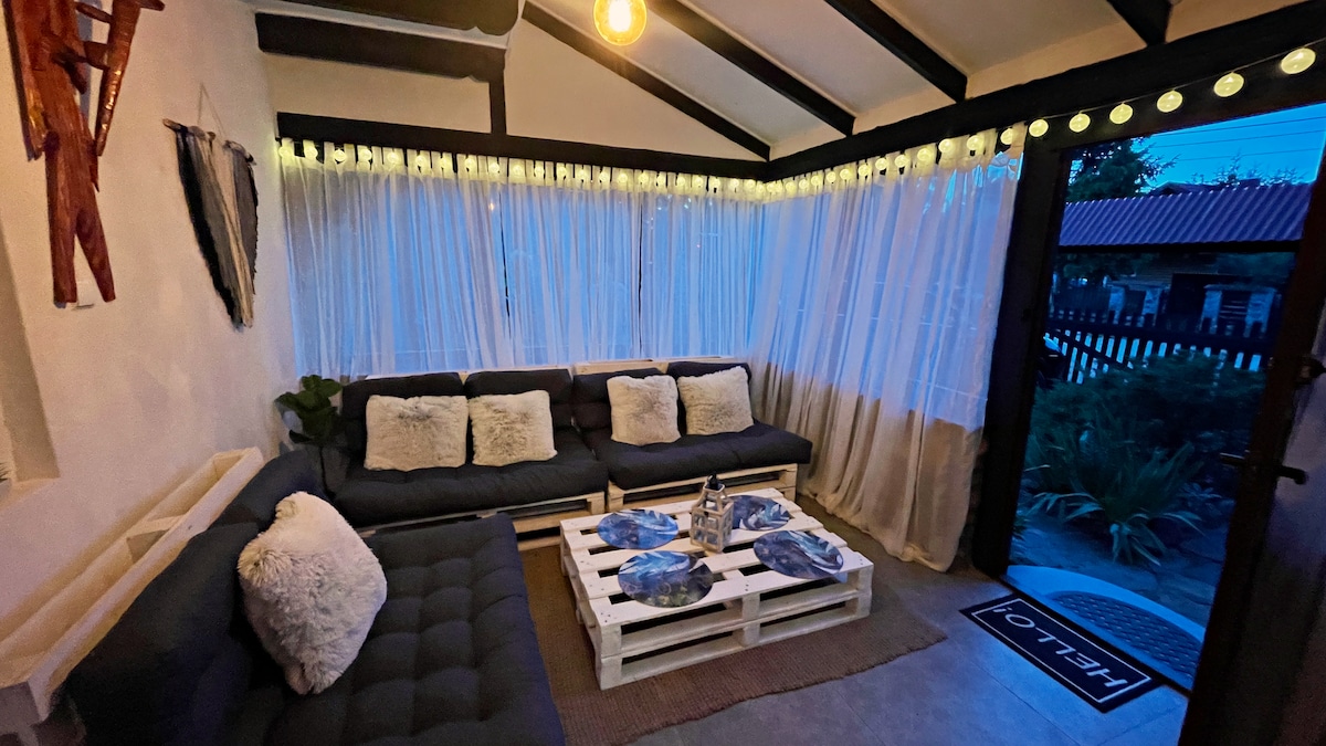 A cozy seating area is featured, furnished with a large sectional sofa adorned with soft cushions. White curtains filter gentle light, and a table set with decorative plates sits at the center. String lights create a warm ambiance, enhancing the relaxing atmosphere.