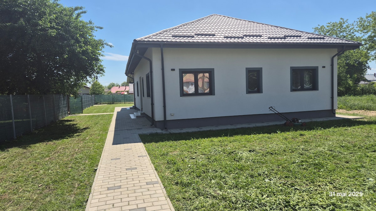 A single-story house is shown, surrounded by a grassy area. The exterior features bright white walls and a sloping roof covered in dark tiles. Multiple windows allow natural light to enter. A paved pathway leading to the entrance is visible, with a fence bordering the property.