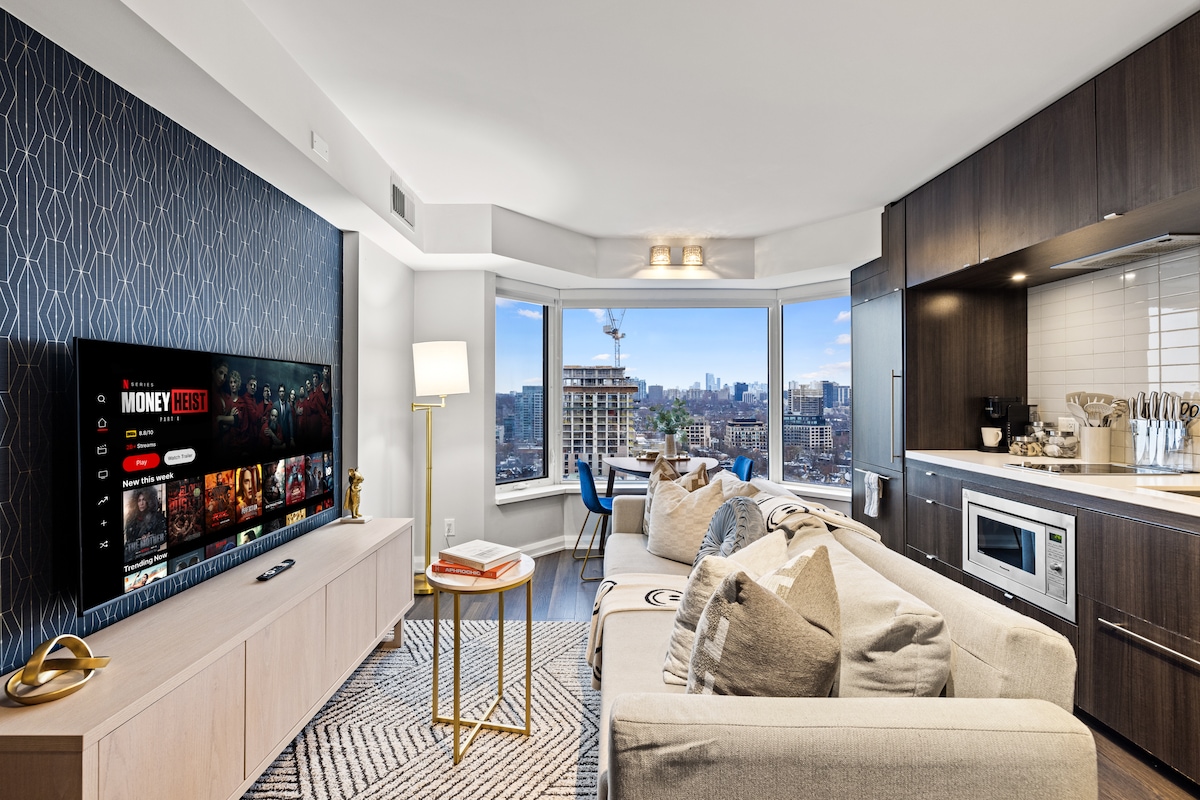 A modern living area is showcased with a large television mounted on a stylish patterned wall. A comfortable sectional sofa is arranged around a coffee table, with city views visible through expansive windows. Warm lighting fixtures add a touch of ambience to the space.