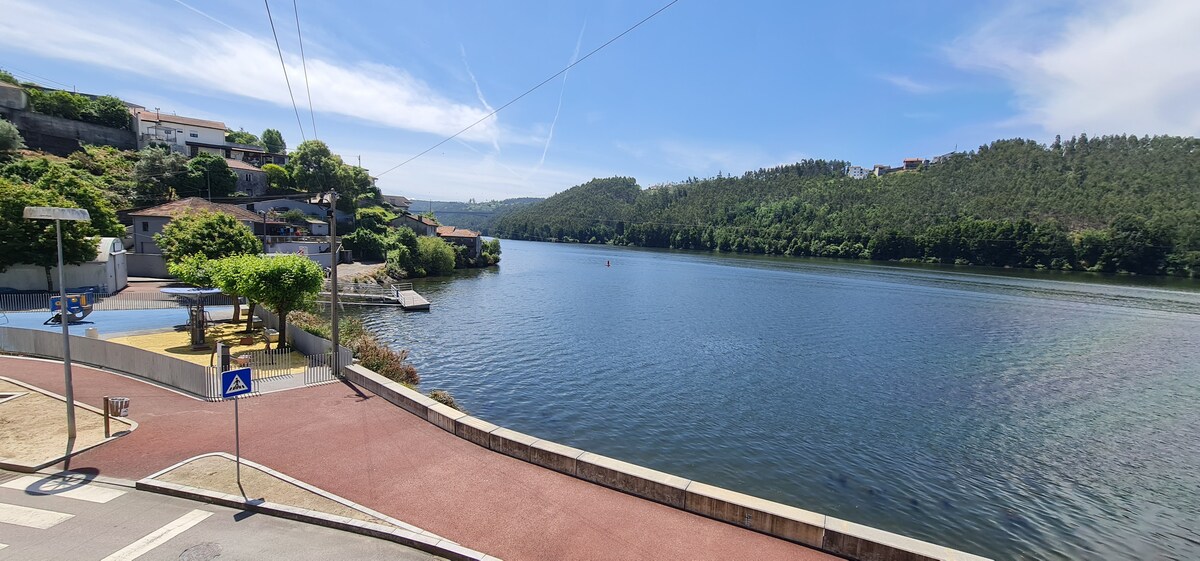 A peaceful view of the Douro River is presented, showcasing gently flowing water bordered by lush greenery on both sides. Clear blue skies reflect the serenity of the landscape, while boats can be seen in the distance, contributing to the tranquil atmosphere.