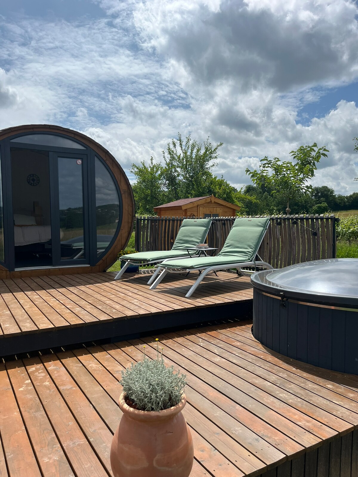 A wooden terrace features two green lounge chairs positioned beside a circular hot tub. The area is embraced by nature, with shrubs and trees visible in the background. The clouds create a dynamic sky above, enhancing the serene outdoor setting.