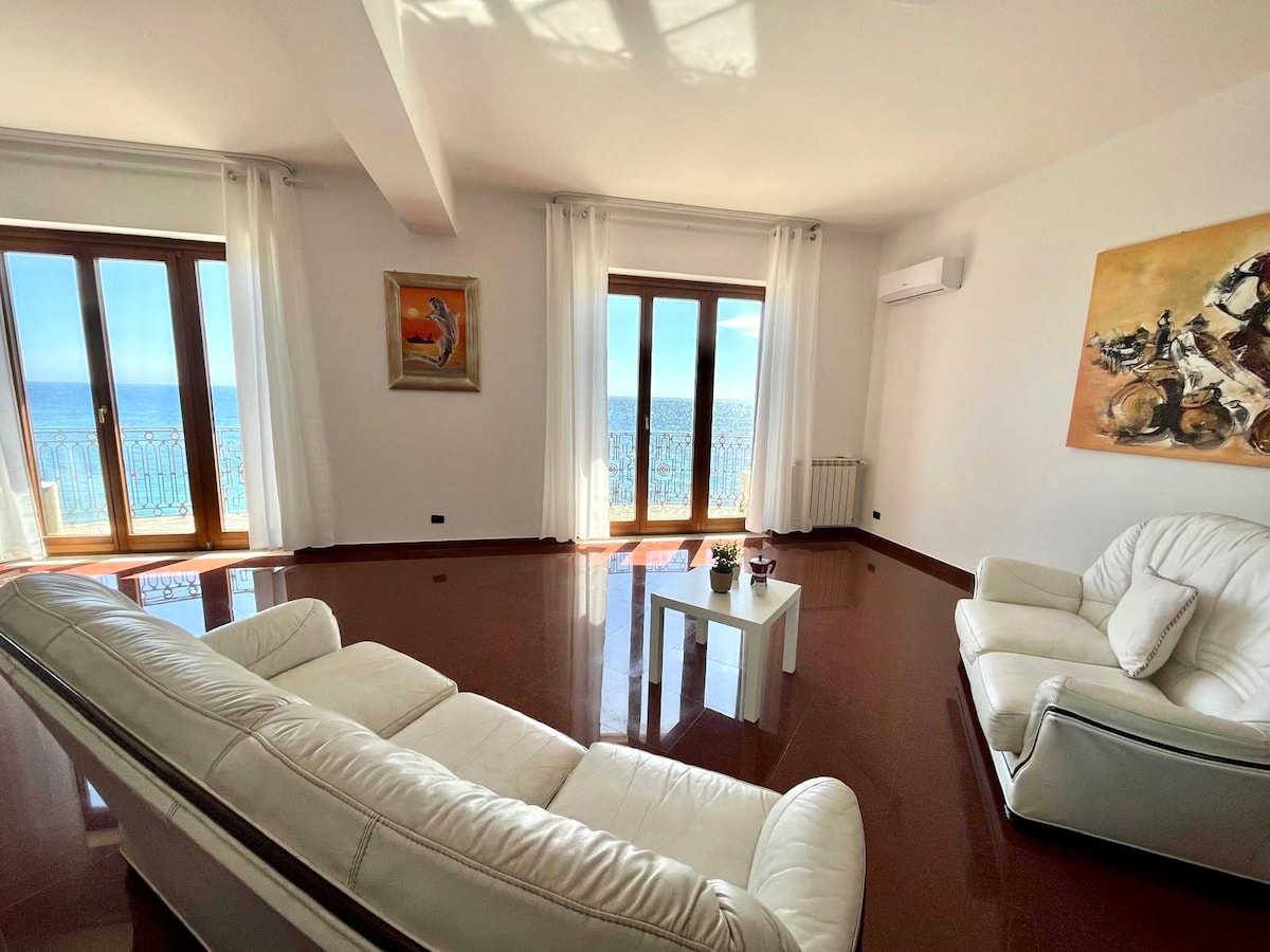 A bright living room features two comfortable leather sofas facing a small white coffee table. Large windows allow natural light to fill the space, showcasing beautiful sea views. Soft curtains frame the windows, and artwork adorns the walls, adding to the serene atmosphere.