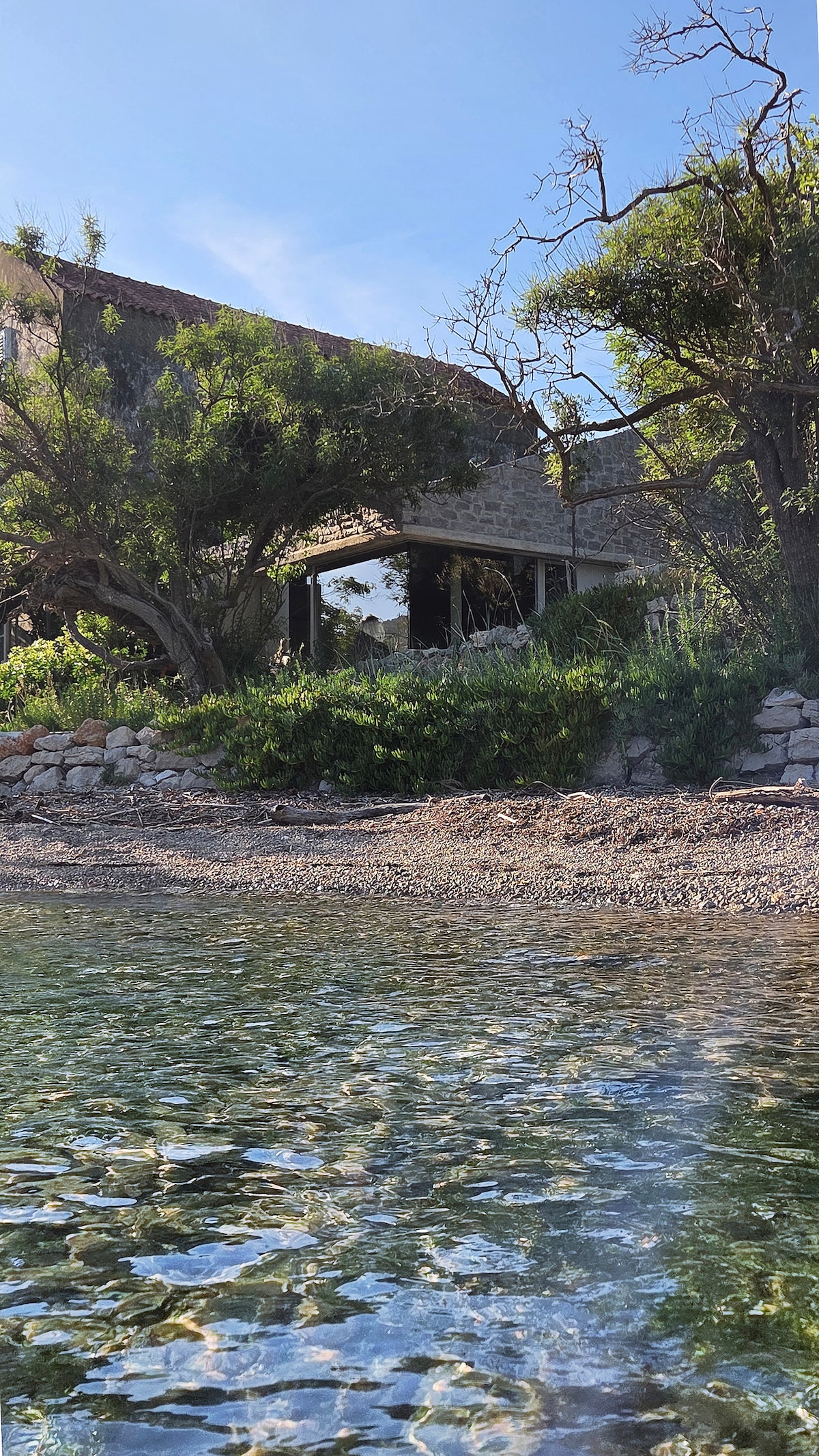 The image captures the exterior of Pretpeć, set against a clear blue sky. Natural stone walls blend with lush greenery, including trees and shrubs, which frame the house. Clear water reflects the sunlight, enhancing the serene beach atmosphere just steps from the house.