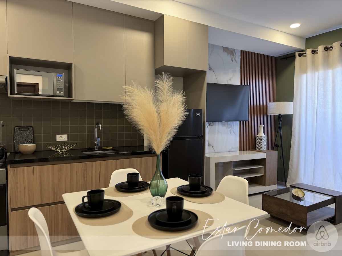 A functional living-dining area is presented with a modern kitchen featuring wooden cabinetry and dark countertops. A white dining table is set with four black plates and cups. Soft neutral tones are complemented by touches of greenery from a decorative plant.