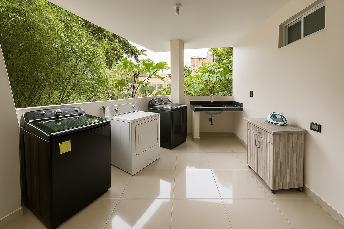 A well-equipped laundry area is visible, featuring a black top-loading washing machine, a white washing machine, and a dark cabinet. A utility sink is positioned alongside a small cabinet with an iron. Large windows provide natural light and views of greenery outside.