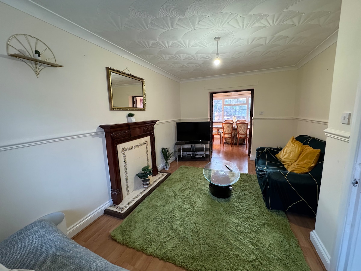 A living area features a green area rug and a glass coffee table at the center. A dark couch is positioned beside a fireplace, with a decorative mirror mounted on the wall. Natural light enters from a window leading to a dining space in the background.
