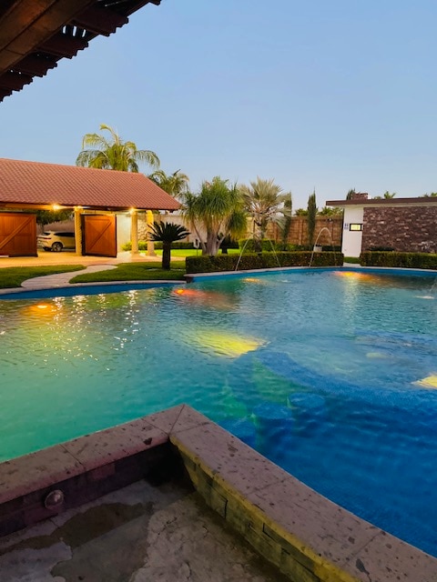 A large swimming pool is surrounded by vibrant greenery and illuminated by colorful lights. The backyard features palm trees and is complemented by well-maintained landscaping. A charming structure with wooden doors is visible in the background, adding to the serene setting.