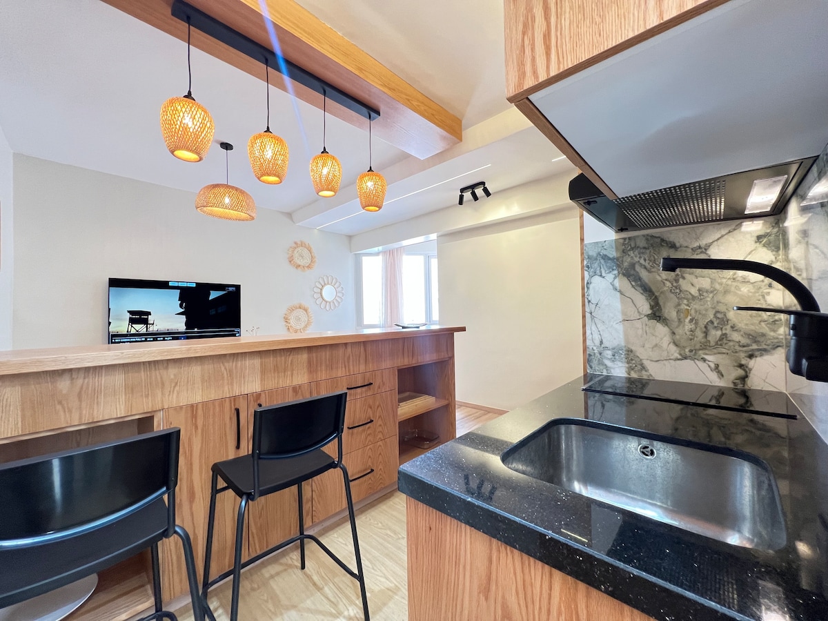 An open kitchen area features a dark countertop with a sink and a modern cooktop. Two black bar stools are positioned near a wooden cooking island, accented by three pendant lights. A television and a round mirror are visible, enhancing the functional space.