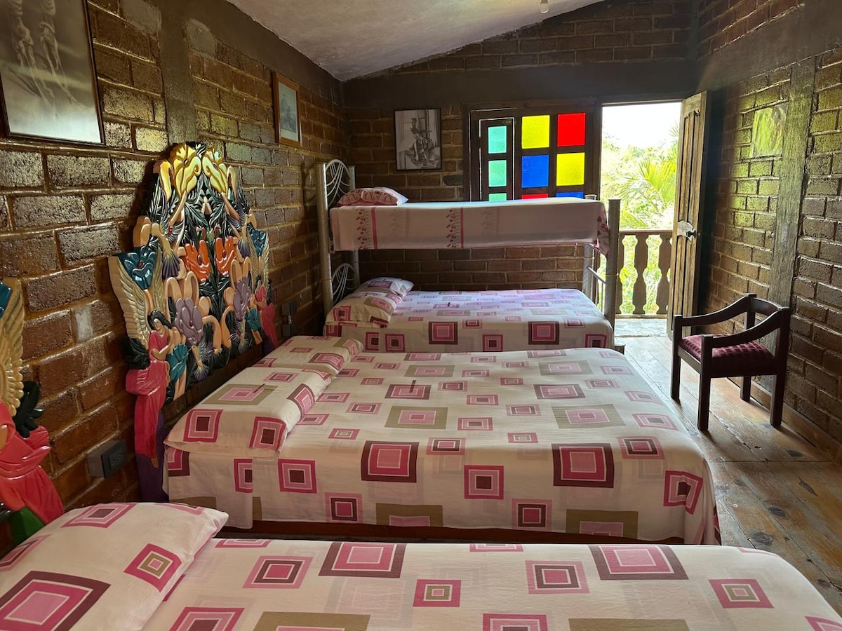 A spacious room adorned with rustic brick walls features three beds, each topped with patterned bedding. A colorful stained-glass window adds a touch of vibrancy, while a wooden chair and a balcony with a view of nature are visible in the background.