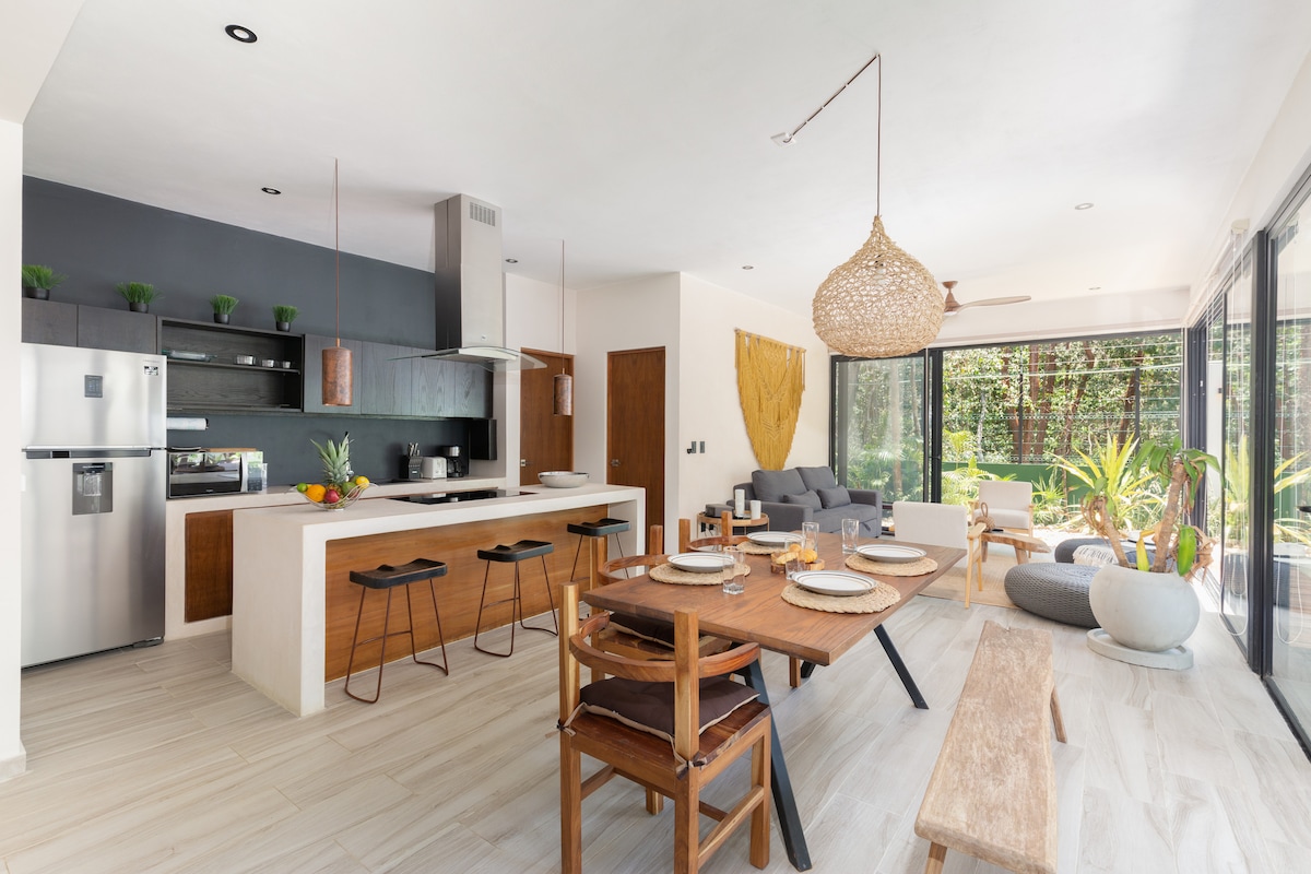 An open-concept living area features a modern kitchen with sleek appliances and a large dining table accompanied by wooden chairs. Natural light floods the space through expansive glass doors, revealing lush greenery outside, while a cozy seating area with a sofa invites relaxation.