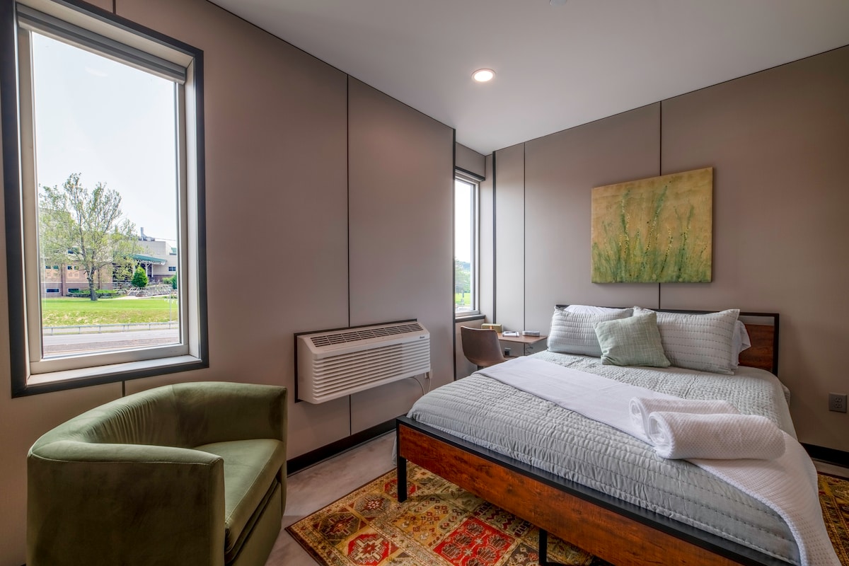 The bedroom features a spacious bed with a light cover, accompanied by a soft, green lounge chair in one corner. Two windows allow natural light to fill the room, while a piece of floral wall art adds a touch of color.