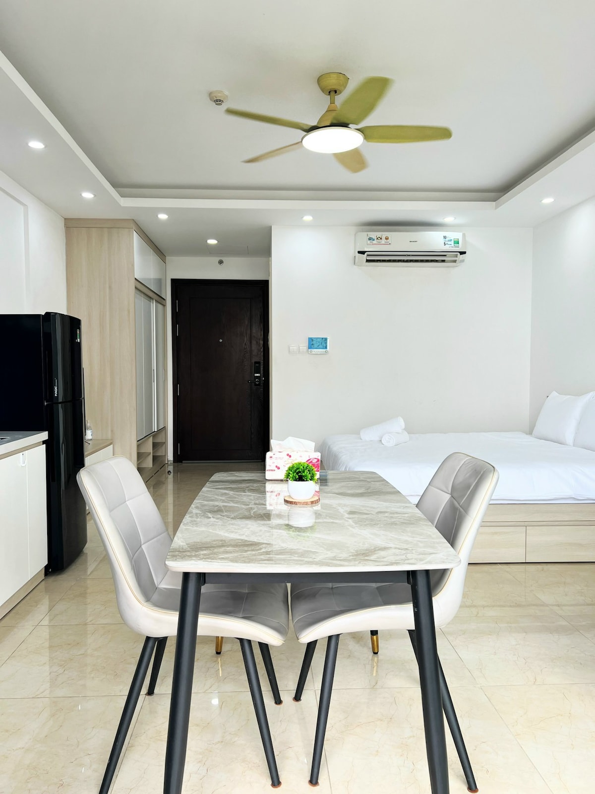 A modern dining area is shown with a marble-topped table and four chairs. In the background, a cozy sleeping area features a spacious bed and a ceiling fan overhead. A kitchenette can be partially seen to the left, while a door leads to the outside.