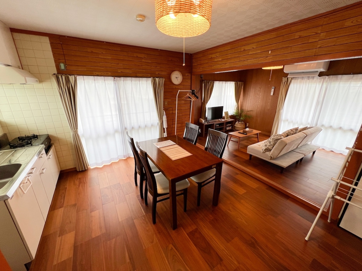 A dining area features a wooden table with four chairs, surrounded by light-colored curtains that diffuse natural light. A cozy living space is visible in the background, equipped with a television and a sofa, enhancing the sense of openness.