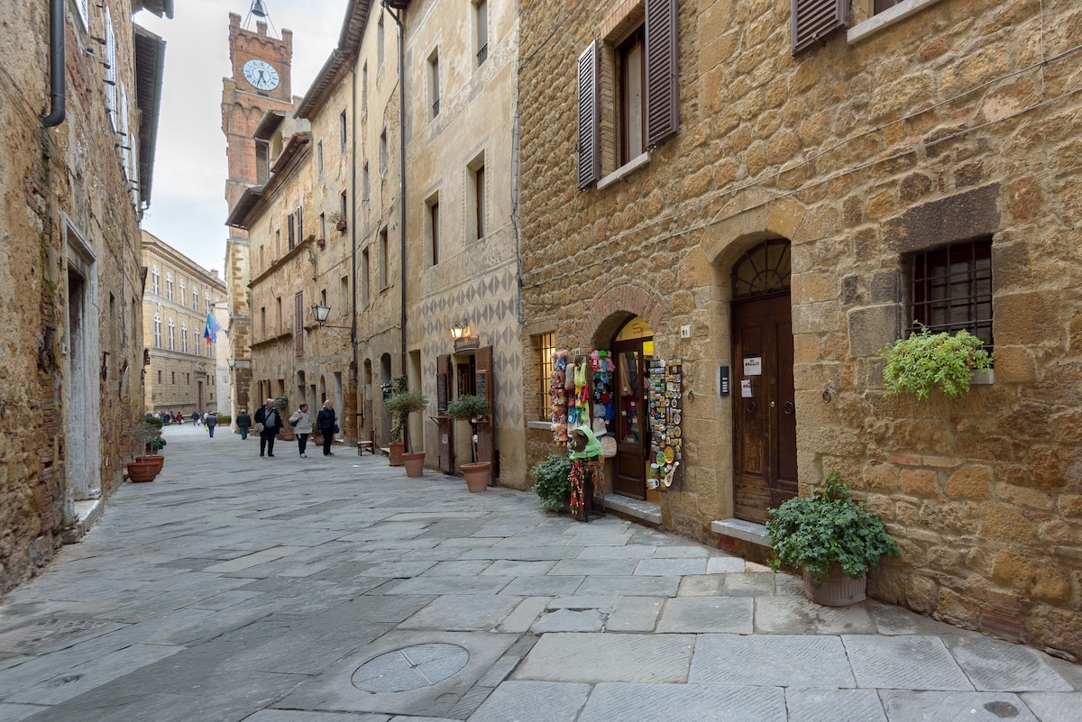 A historic street in Pienza features charming stone buildings with wooden shutters and small shops showcasing vibrant displays. Potted plants line the walkways, while people stroll leisurely along the cobblestone path. The clock tower can be seen in the background, adding to the scenic atmosphere.