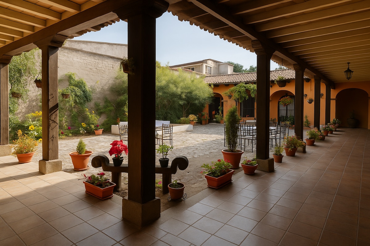 A wide corridor framed by wooden columns opens to a serene central courtyard. Potted plants line the tiled floor, enhancing the calming environment. A stone pathway leads to an area with chairs and tables, surrounded by greenery, creating a tranquil space for relaxation.