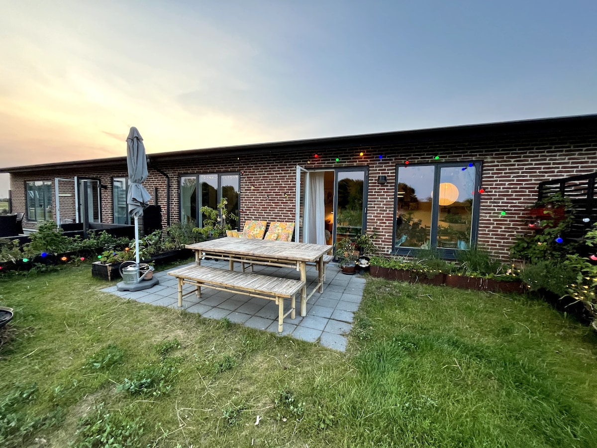 A cozy outdoor seating area is highlighted with a wooden table and benches, surrounded by green grass and potted plants. Large glass doors offer access to an interior space, with colorful decorative lights adding a festive touch as the sun sets in the background.