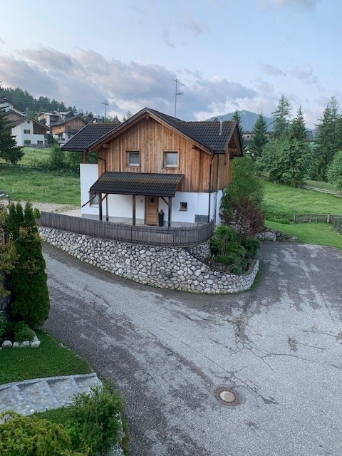 A charming alpine chalet is positioned on a gently sloping lot, featuring a combination of wooden and white facades. Surrounding greenery enhances the tranquil environment, while a circular driveway with stone accents leads to the main entrance, showcasing a welcoming facade.