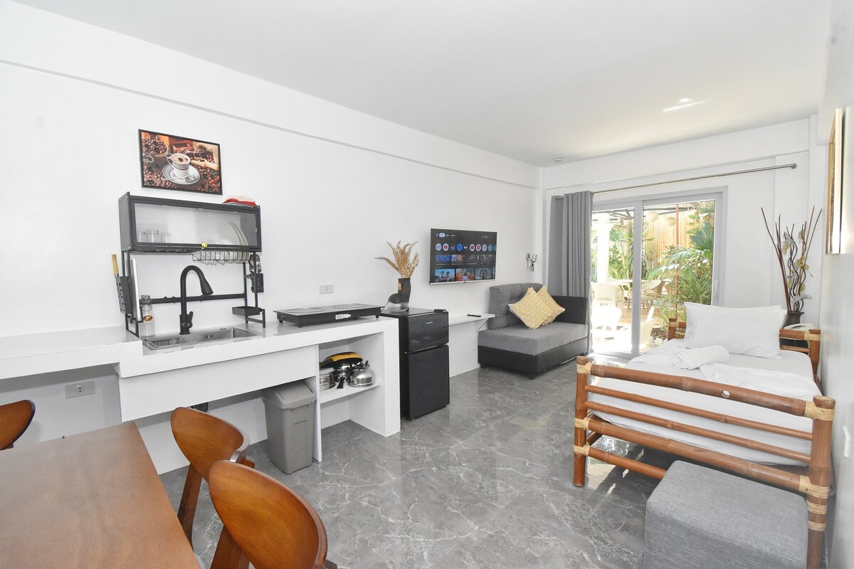 A bright and open living space is shown, featuring a dining area with wooden chairs, a modern kitchenette equipped with appliances, and a cozy sofa. Natural light enters through glass doors leading to an outdoor area, with decorative elements and a Smart TV enhancing the room's functionality.