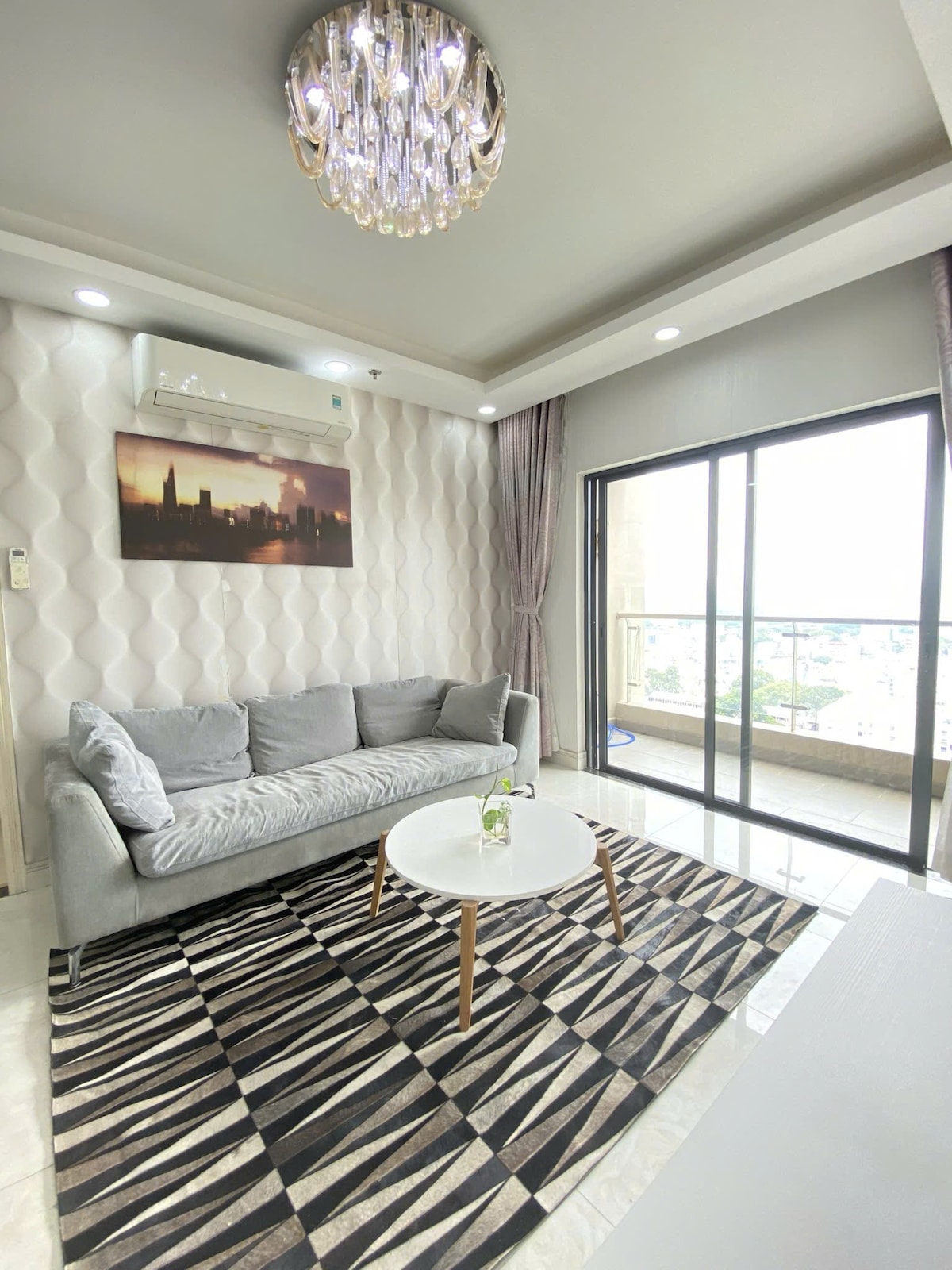 A modern living area showcases a light gray sofa centered on a patterned area rug. A circular coffee table stands in front, accompanied by a delicate floral arrangement. Natural light enters through sliding glass doors, with city views visible beyond.