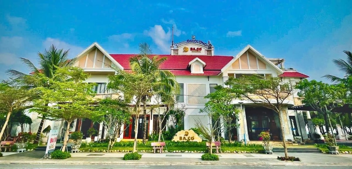 The hotel exterior showcases a charming architectural design with a red roof and light-colored walls. Lush greenery surrounds the entrance, complemented by palm trees and well-maintained landscaping. Benches are positioned along the walkway, enhancing the inviting atmosphere of the space.