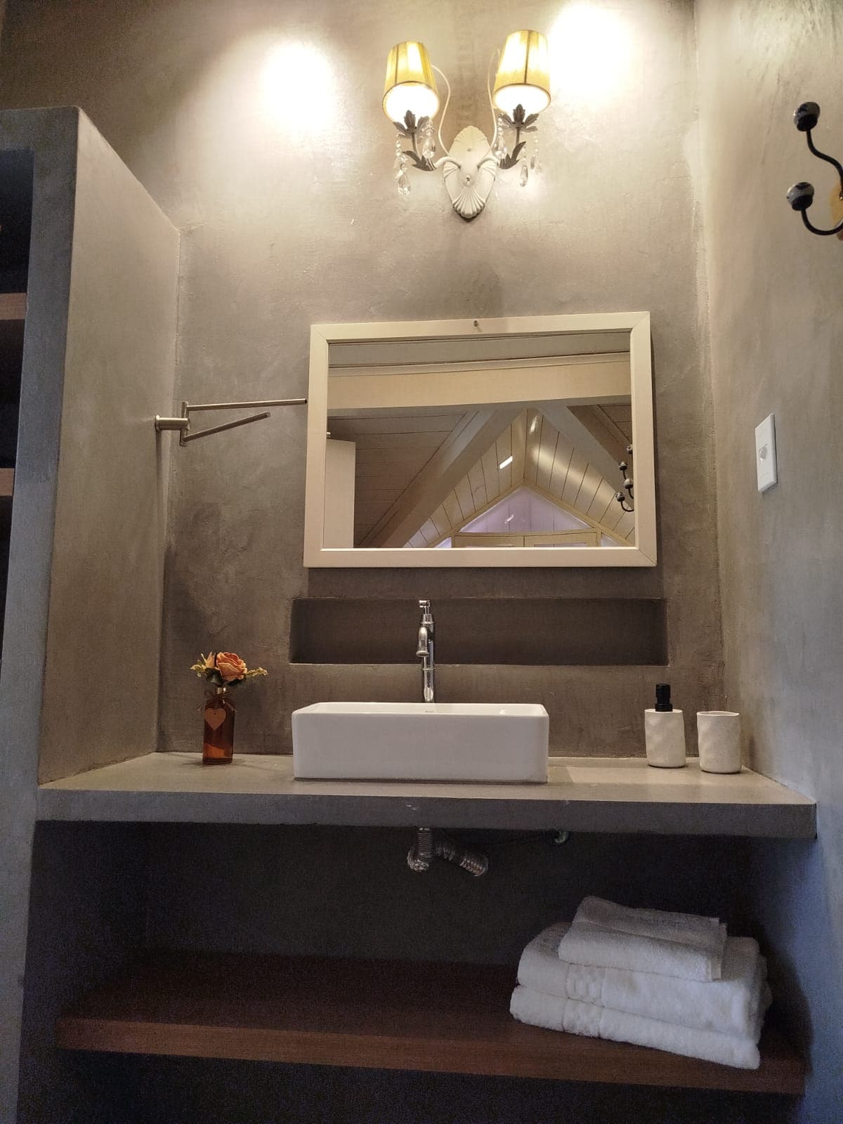 A minimalist bathroom area features a sleek, square sink positioned on a concrete countertop. A wall-mounted mirror reflects the room's light fixtures. Fresh flowers in a small vase and neatly arranged toiletries add a touch of detail. Soft towels are stacked on a lower wooden shelf.
