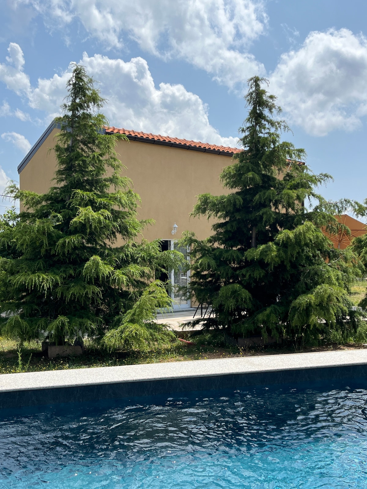 A serene outdoor view showcases a modest two-story home framed by lush evergreen trees. In the foreground, a calm pool reflects the clear blue sky and puffy clouds, creating a tranquil setting perfect for relaxation.