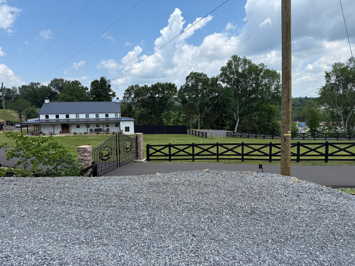 A well-maintained gravel driveway leads to a white barn-style residence, surrounded by lush greenery and tall trees. A black wooden fence encloses an expansive, manicured lawn, providing an open view of the serene landscape under a partly cloudy sky.