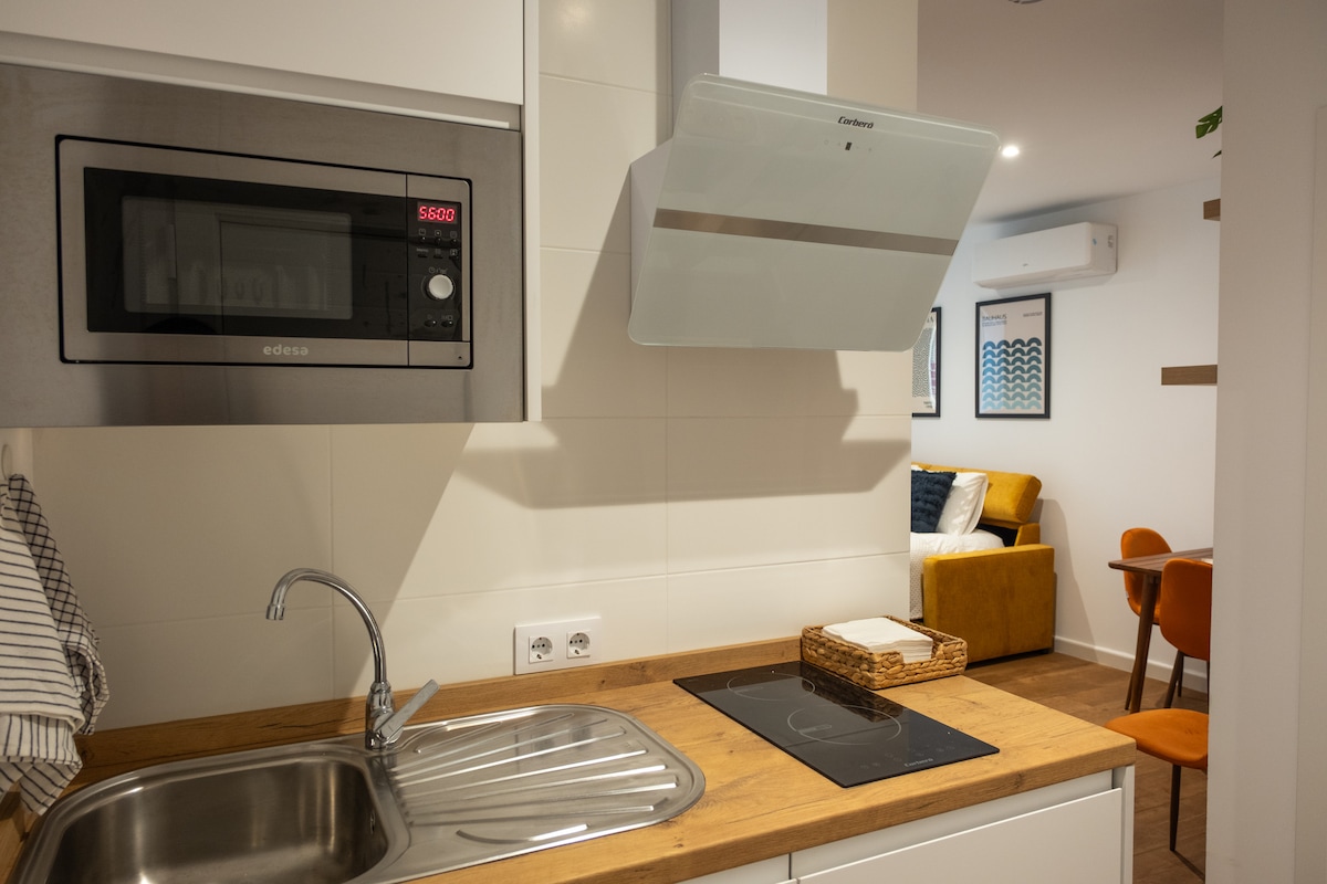 A modern kitchen is presented, featuring a stainless steel sink and an induction cooktop surrounded by sleek cabinetry. A microwave is built into the wall above the counter. Warm wooden surfaces add a touch of coziness, while a glimpse of the living area is visible in the background.