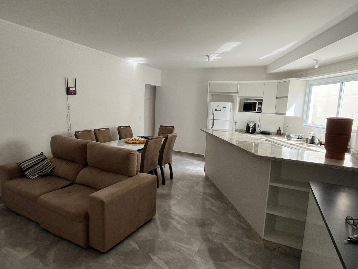 An open-concept living area features a beige sectional sofa surrounding a cozy coffee table. A dining table with several chairs is positioned nearby, while a kitchen area showcases modern cabinetry and appliances. Natural light enters through a window, illuminating the space.