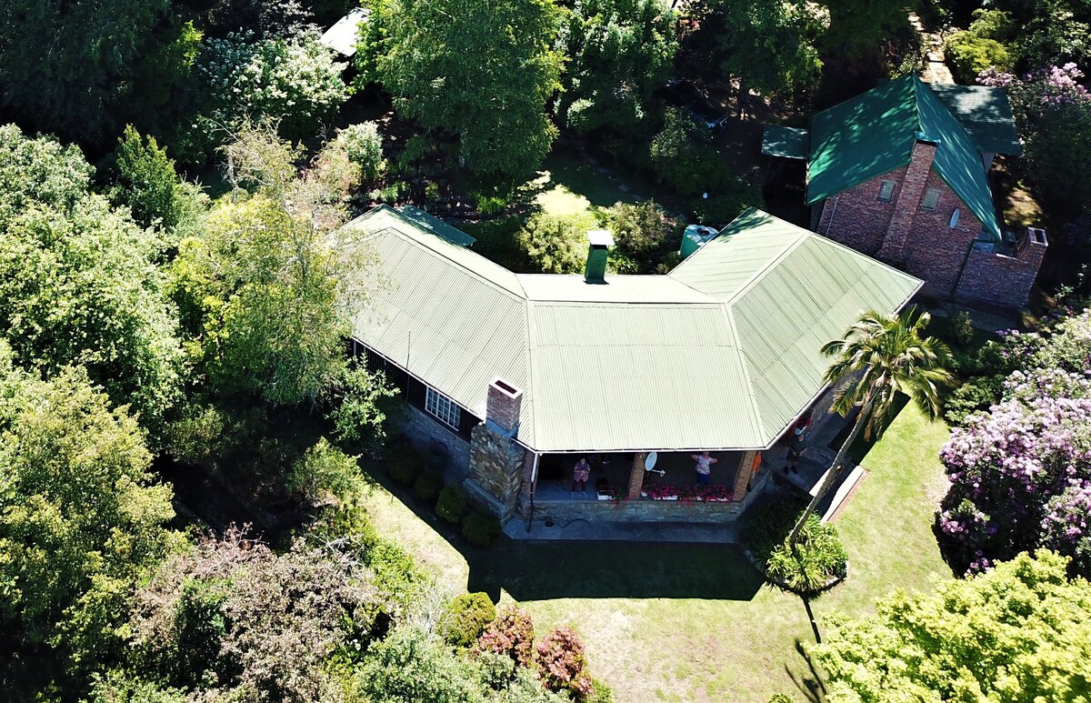 An aerial view showcases a spacious dwelling surrounded by lush greenery and colorful flowering plants. The structure features a characteristic green roof and is positioned within a tranquil garden space, offering a peaceful ambiance. Trees and manicured shrubs frame the property's perimeter.