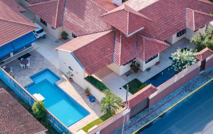 An aerial view of a spacious property is presented, showcasing a well-maintained swimming pool adjacent to a patio area, surrounded by landscaped gardens. The roof features a classic red tile design, complementing the light-colored exterior walls of the house.