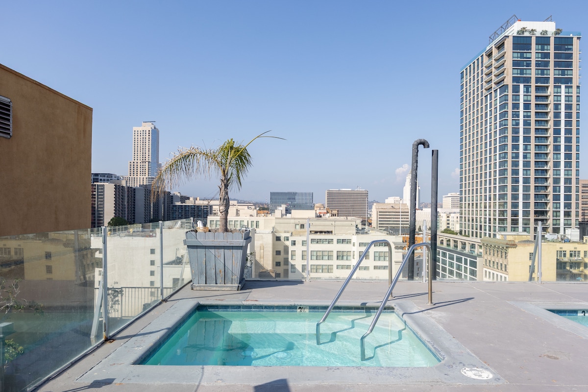 A rooftop pool area is visible, featuring a clear view of the skyline with modern buildings in the background. A palm tree adds greenery to the space, while a metal ladder leads into the pool. Glass railings provide safety and unobstructed views.