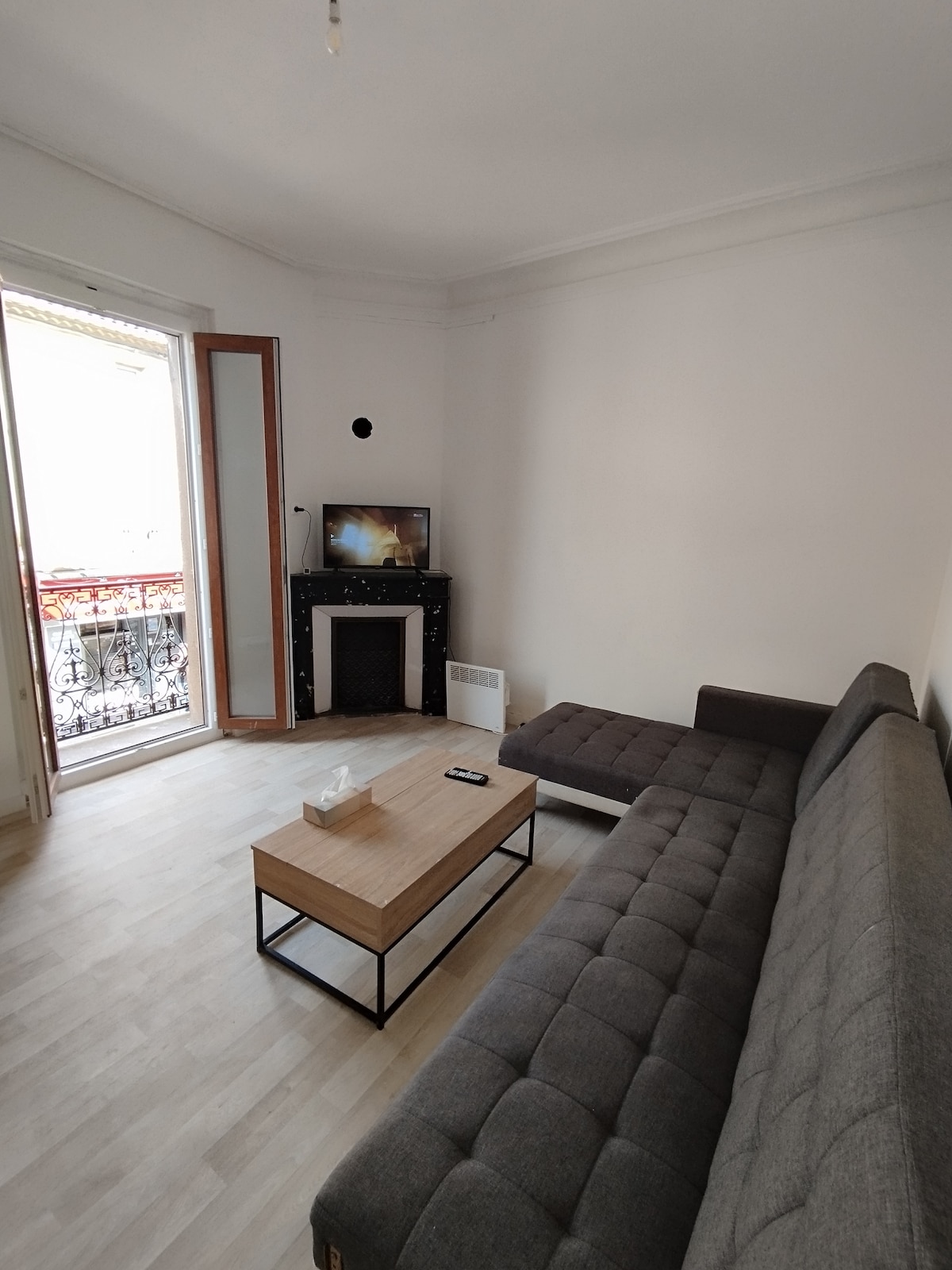 A cozy living area is highlighted by a neutral color palette. A functional coffee table sits in front of a comfortable sectional sofa. A vintage fireplace adds character, while a TV is positioned for easy viewing. Natural light filters through the open balcony doors.