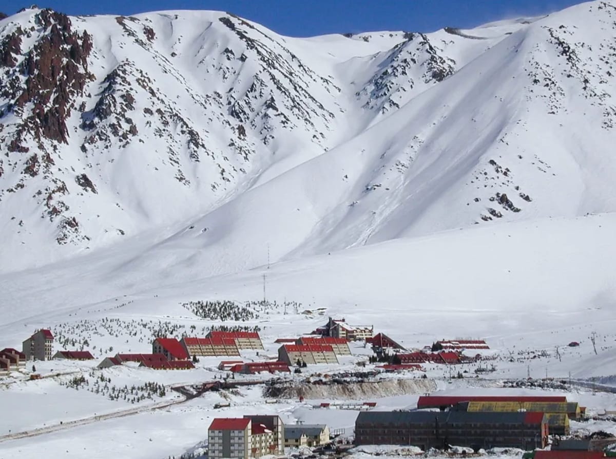 A scenic view of snow-covered mountains frames a series of buildings below, featuring red roofs against a backdrop of white slopes. The expansive landscape highlights a serene winter setting, with gently rolling hills and open spaces partially obscured by fresh snowfall.