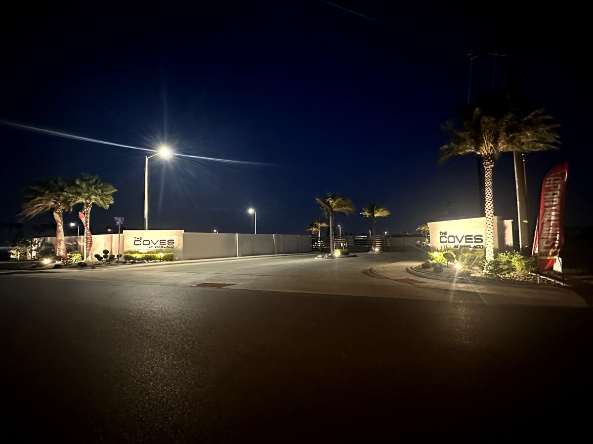 The entrance to a gated community is captured at night, featuring illuminated signage that reads 'Coves.' Palm trees line the entrance, and streetlights provide ample lighting along the smooth road, creating a welcoming approach to the community.