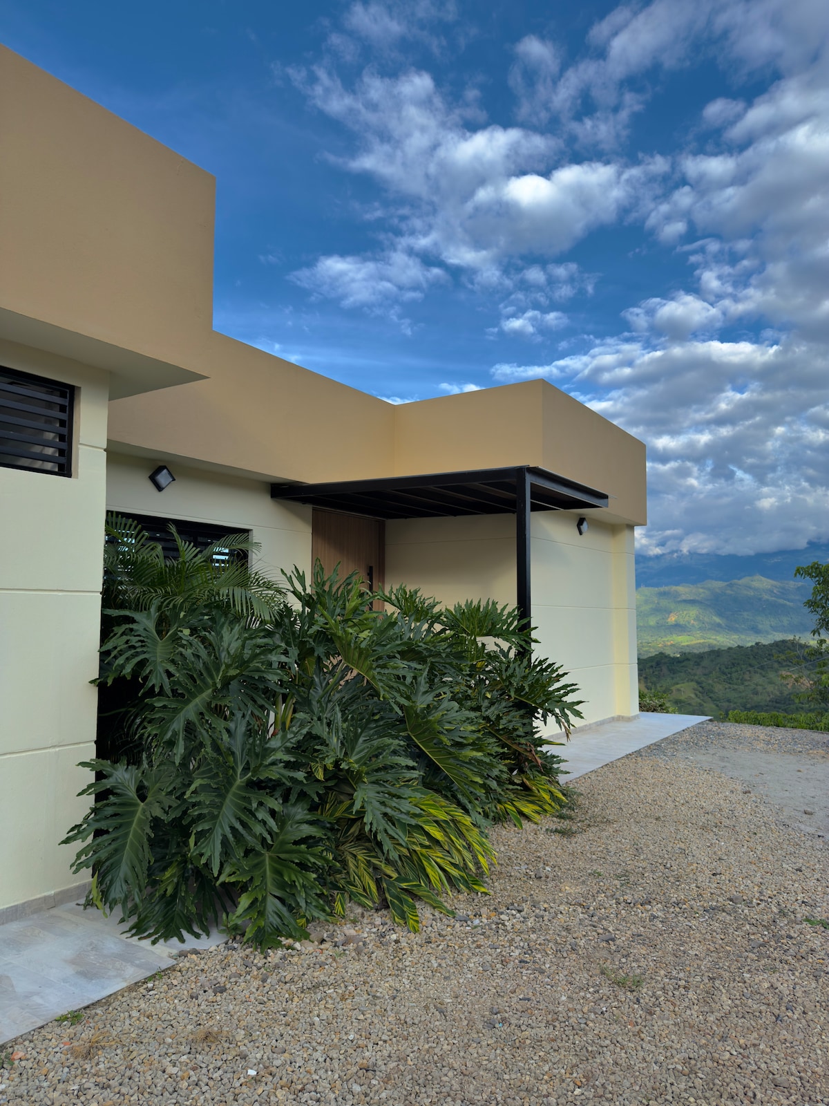 A modern structure is nestled among lush greenery, with large leaves framing the entrance. The building features clean lines and a flat roof, while a gravel pathway leads to the doorway. A panoramic view of mountains is visible in the background under a partly cloudy sky.