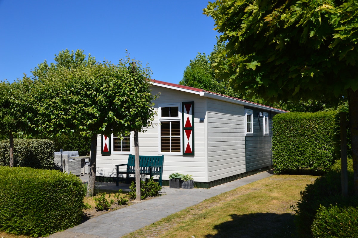 A modern chalet is surrounded by well-maintained hedges and trees, with a clear blue sky above. Two bright red and white window shutters contrast with the light-colored exterior, while a small patio area with seating is visible to the side.