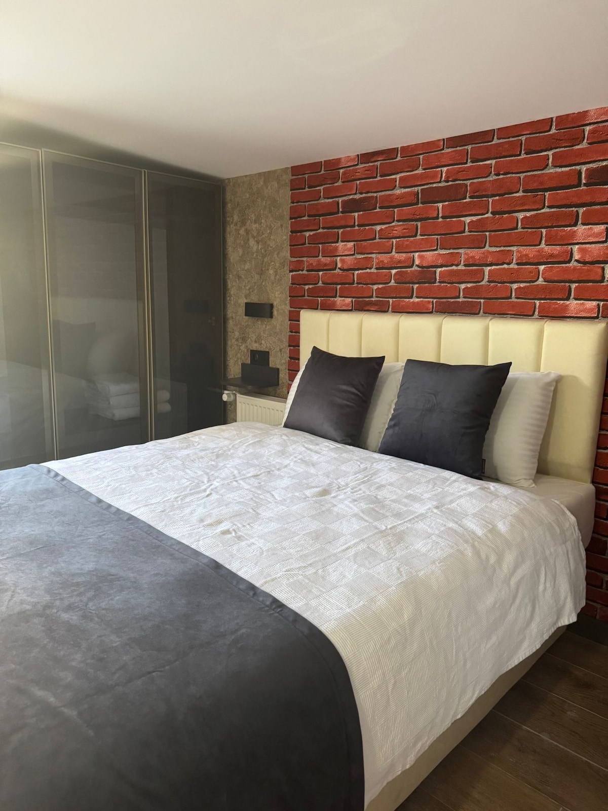 A spacious bed is layered with a light quilt and decorative pillows in shades of gray and black. Behind the bed, a brick accent wall adds a contrasting texture. Soft lighting enhances the calming ambiance, complemented by a wardrobe visible in the background.