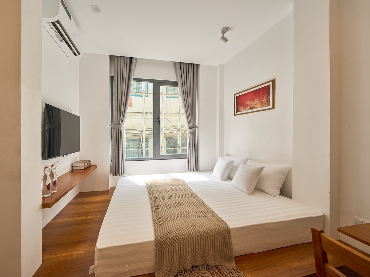 A comfortable sleeping area features a spacious bed with a light blanket draped across it. Two pillows are placed at the head of the bed. Natural light enters through large windows adorned with soft curtains, while a mounted television is positioned on the opposite wall.