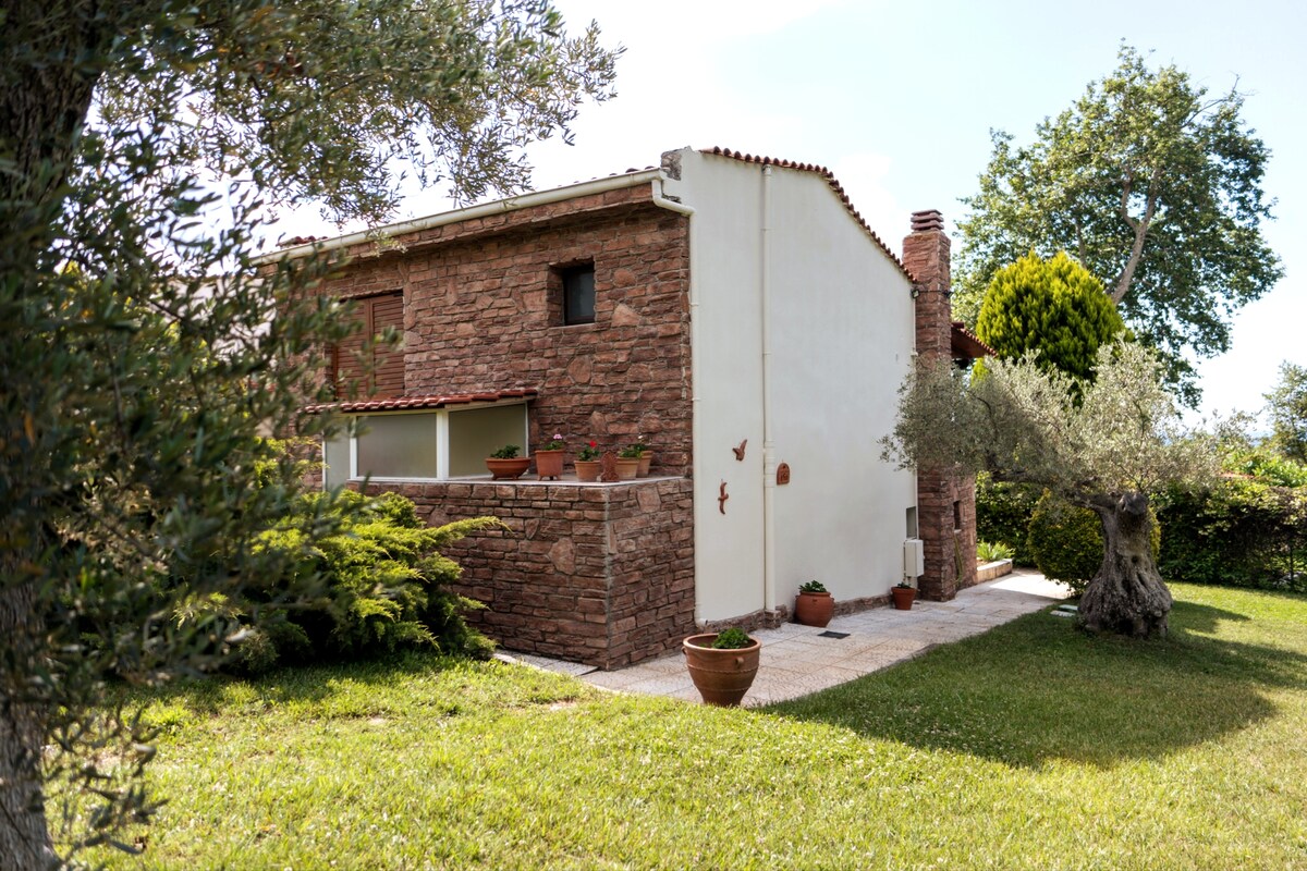 A rustic stone house is situated in a green landscape, surrounded by trees and grass. The structure features a combination of natural materials, with potted plants adorning the exterior. A pathway leads to the entrance, creating a welcoming appeal in a serene environment.
