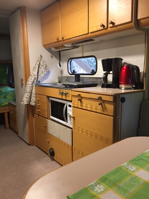 A compact kitchenette features wooden cabinetry with multiple storage compartments. A microwave and coffee maker are present on the counter beside a red kettle. A colorful tablecloth is set on a nearby table, while a window allows natural light to brighten the space.