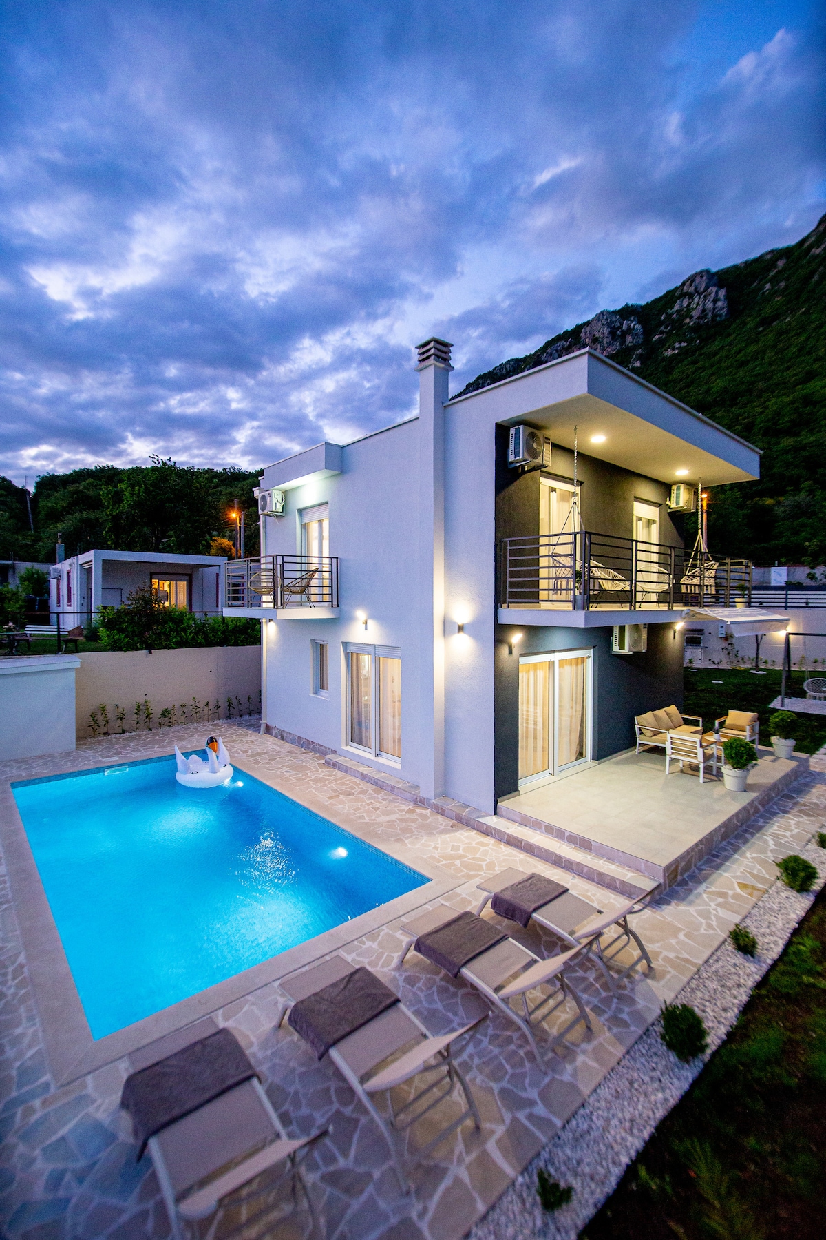 A modern two-story villa is situated beside a private swimming pool, surrounded by patio chairs. The building features large windows and balconies, with a backdrop of lush greenery and distant mountains under a twilight sky.