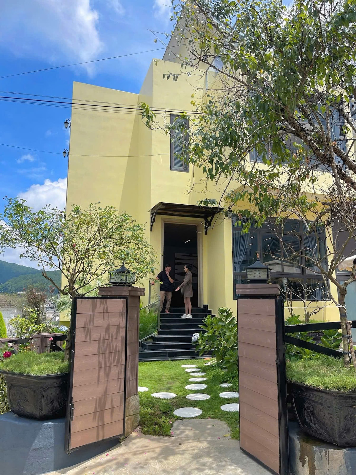 The entrance to the villa is framed by a well-maintained garden, featuring a pathway of stepping stones leading to the front door. Soft greenery and decorative planters are positioned on either side of the entrance. Large windows and a yellow exterior create an inviting atmosphere.